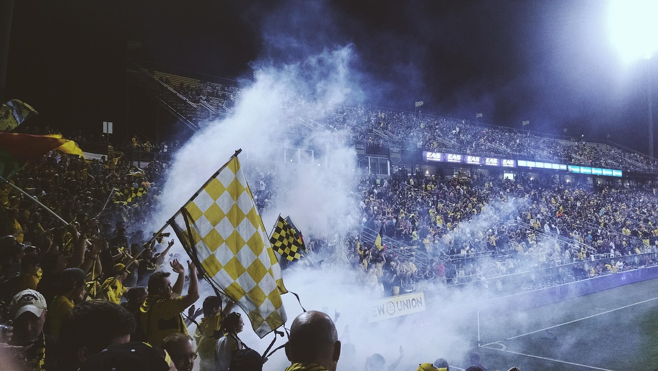 Des supporters agitent des drapeaux dans une tribune enfumée, sous les projecteurs d’un stade