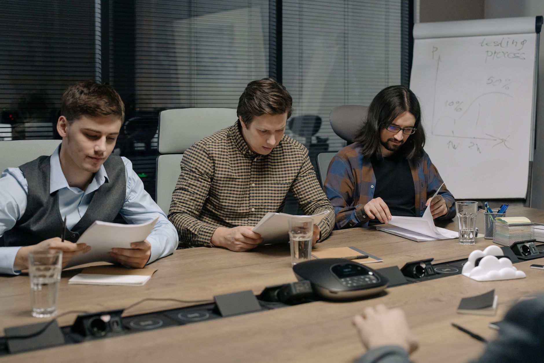 Trois hommes assis à une table de réunion lisent des feuilles, avec un téléphone de conférence et un tableau à feuilles derrière eux.