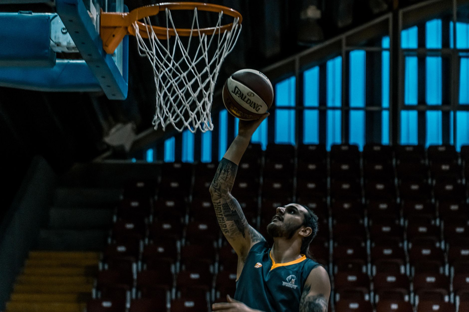 Un joueur de basket tatoué, en maillot sombre, monte vers le panier avec un ballon dans une salle aux gradins vides.