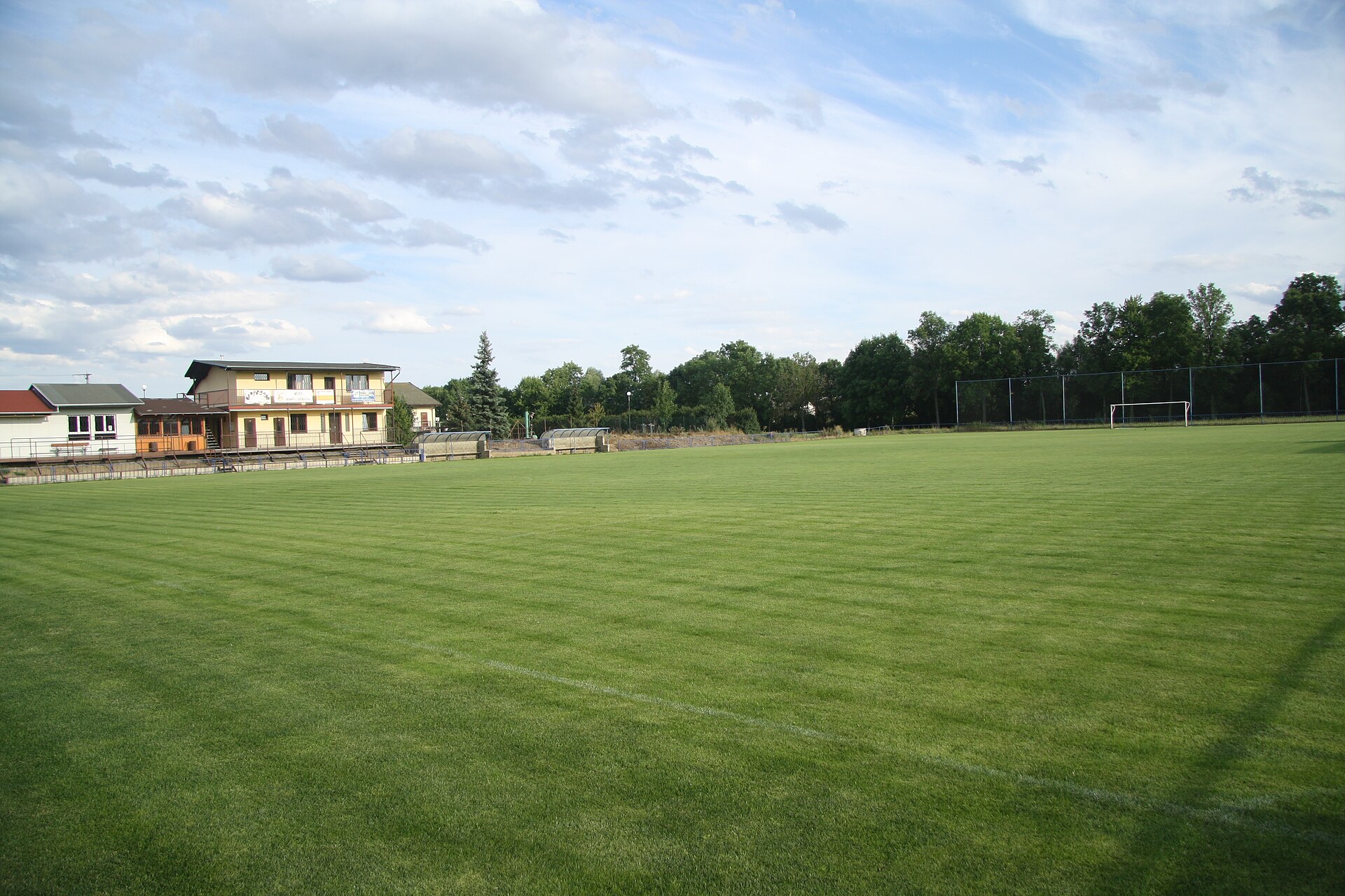 Vaste pelouse de football tondue en bandes, avec un but au loin, un petit club-house et des arbres sous un ciel nuageux